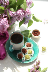 Obraz na płótnie Canvas On a white background is a cup with a coffee, on a turquoise plate of candy and truffle, and in a vase is a lilac. 
