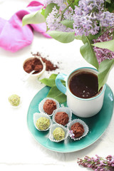 On a white background is a cup with a coffee, on a turquoise plate of candy and truffle, and in a vase is a lilac.
