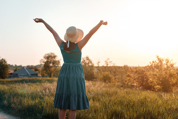 happy young woman at summer sunset in the field raises her hands to the sky