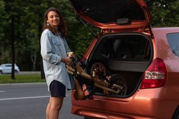 A young woman in casual clothes takes a folding bike out of the trunk of a car. Lays in the trunk of an orange car in the evening.