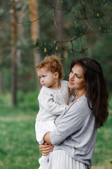 young attractive girl with long hair, brunette, in clothes of neutral colors, stands in the summer forest with her daughter little baby girl