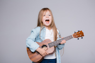 Fototapeta premium The child plays on the ukulele and sings songs. Joyful girl stands in a blue shirt on a gray background