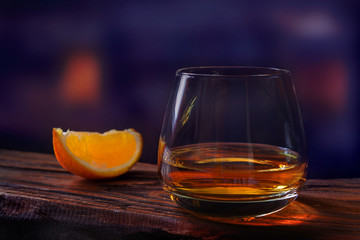 Whiskey drink glass with orange slice on wooden table on dark blurred bar background