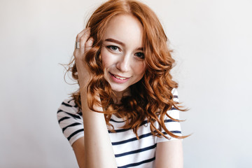 Fototapeta premium Wonderful dark-eyed girl touching her ginger hair with gently smile. Indoor photo of positive european lady in striped t-shirt.