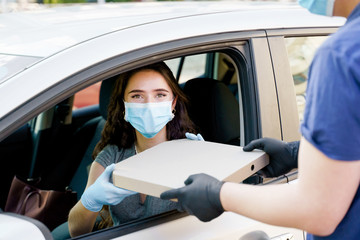 Food delivery courier gives pizza to woman in eco car. Safety food delivered according social distance. Tasty pizza with salami, cheese, tomatoes in cardoard box. Giving food trought car`s window.