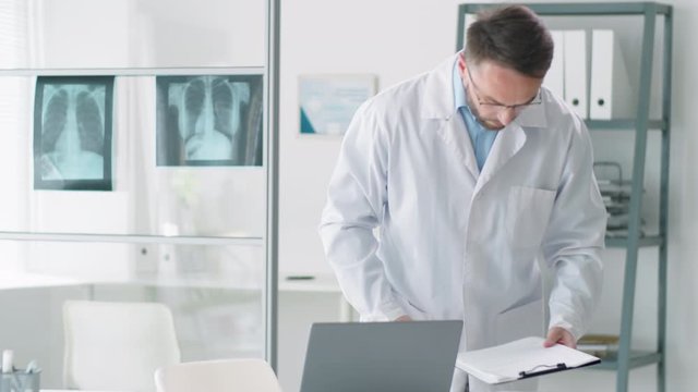 Professional male doctor in lab coat holding clipboard, walking into medical office, sitting at desk and typing on laptop during workday