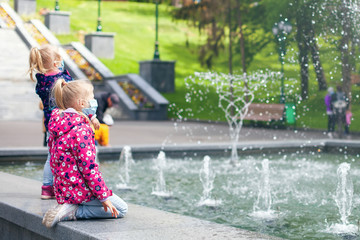 Children walk in the park near the fountain on a bad sunny day. Medical masks on faces, good mood. The child holds a finger up. quarantine weakening