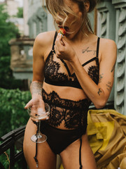 Young beautiful girl in black lingerie and a beige trench coat drinks champagne on a balcony in the...