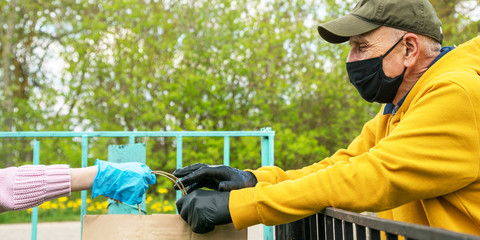 girl hand in blue glove gives paper bag to old man by fence