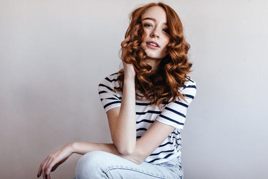 Interested ginger woman in jeans sitting in studio and looking to camera. Indoor photo of good-humoured red-haired girl in trendy casual t-shirt.