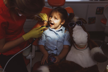 High angle of female doctor using professional instrument to examine teeth of little girl under bright light in dark dentist office