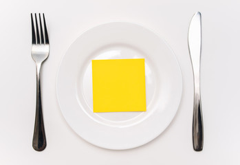A big white plate, knife and fork lie on a tablecloth. Empty sheet of yellow paper for copy space and mockup, top view.