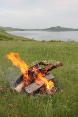 Bonfire in the foreground in a green meadow on the lake