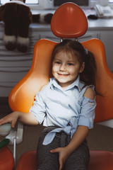 Cheerful little patient smiling and looking at camera while sitting in dentist chair under spot of light and waiting for appointment