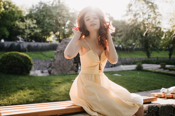 Excited girl in romantic yellow dress sitting on bench in park. Outdoor shot of graceful ginger woman touching her hat in sunny morning.