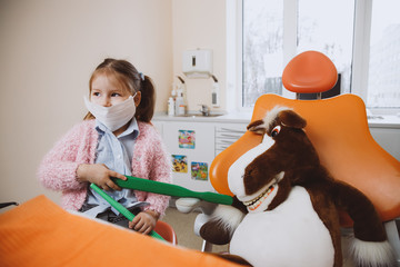 Little girl in medical mask using huge brush to clean teeth of toy and looking away while playing in dentist office in modern clinic