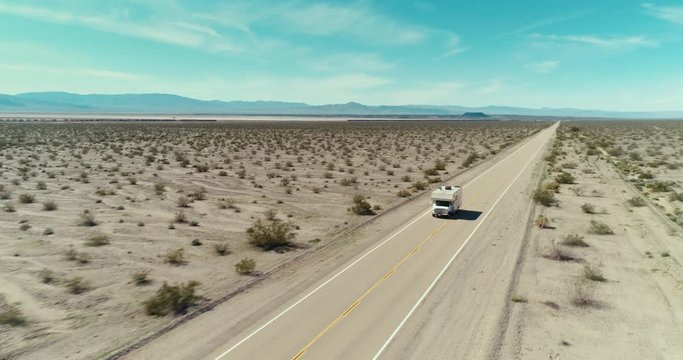 AERIAL - A drone tracks a small brown camper from the front in the middle of the Mojave desert on a long road on a clear and blue day in the summer with mountains in the back