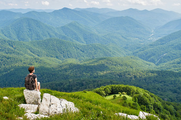 Woman with backpack sitting on top of the mountain. View of the green meadow. Mountain tourism and a sense of freedom.