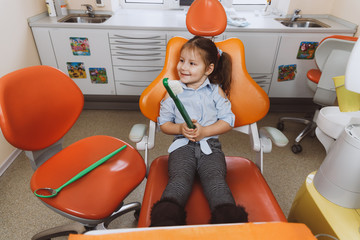 High angle of happy little girl with large toothbrush smiling and looking away while sitting on chair and waiting in dentist office