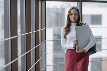 attractive girl walks in a bright corridor with laptop and things in airport terminal or office. travel atmosphere or alternative work atmosphere. freelancer student travels to business meeting.