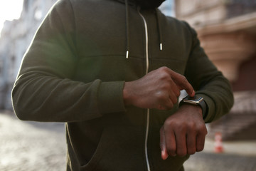 Obraz na płótnie Canvas Using fitness app. Cropped photo of african male runner in black sportswear touching his smartwatch while running outdoors in the morning