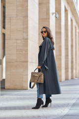 Naklejka premium Full length portrait. Woman walking city street. Female model holding leather reptile handbag. Stylish girl in gray raincoat and sunglasses. Caucasian brunette lady with wavy hair
