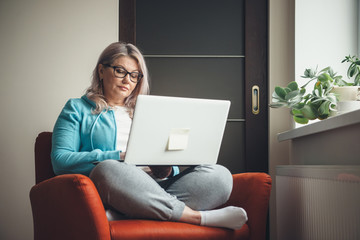 Blonde caucasian aged woman with eyeglasses resting in the armchair at the laptop