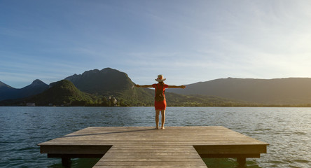 Girl standing up with a stalk hat on and red dress with her arms up looking at the mountains with her shoes on a dock by a lake - Powered by Adobe