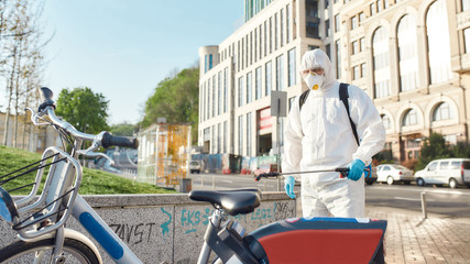 Break the germ cycle. Sanitization, cleaning and disinfection of the city due to the emergence of the Covid19 virus. Man in protective suit and mask at work near rental bike stop