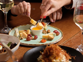 Female hand holding canape near cheese platter with different cheese and grapes on a table for brunch. female friends - hands clinking white wine glasses, restaurant or bar on background, close up