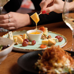 Fototapeta na wymiar Female hand holding canape near cheese platter with different cheese, honey and grapes on a table for brunch. female friends - hands clinking white wine glasses, restaurant or bar on background, close