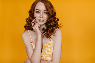 Joyful caucasian girl with ginger hair touching her face with smile. Indoor photo of spectacular young female model in yellow attire