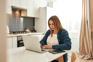 Full concentration. Young focused caucasian woman in casual clothes sitting at kitchen table at home and using laptop