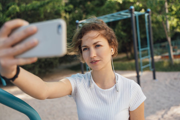 A brunette girl with blue eyes in a white top takes a selfie on a mobile phone with headphones staring at the camera smiling middle plan