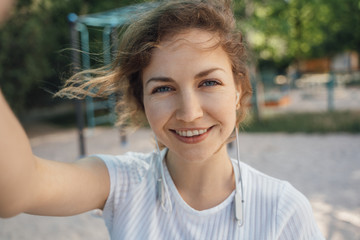 Girl brunette with blue eyes in a white top takes a selfie on a mobile phone with headphones looks into the camera smiling close-up