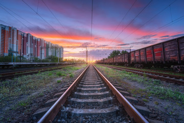 Fototapeta na wymiar Railway station with freight trains and multicolored buildings at sunset. Railroad in summer. Heavy industry. Landscape with train, railway platform, sky with colorful clouds at dusk. Transportation