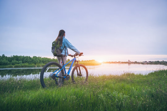 Woman with bicycle on the hill near the river at sunset in spring. Landscape with sporty girl with backpack riding a mountain bike, dirt road, green grass, water in summer. Sport and travel. Cycle