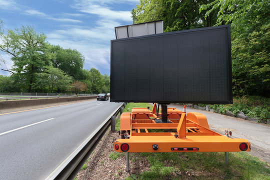 A Blank Digital Highway Shoulder Sign, For Copy Space, With The Road On One Side And A Car Driving Away In The Distance, Surrounded By Foliage. A Blue Sky With Wispy Clouds In The Background.