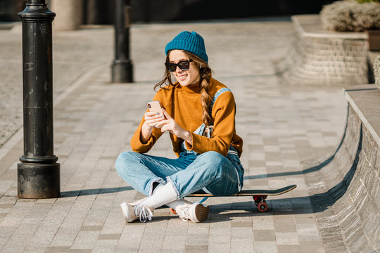 Girl sitting on skateboard and use mobile phone. Outdoors, urban lifestyle. cute skater girl sitting on skate board checking smart phone listening to music using internet and takes a photo - Powered by Adobe