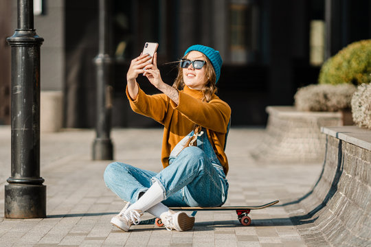 Young Woman Sitting On Skateboard, Using Phone. Skateboarding Woman In City. Female Skate Boarder With Smartphone And Skateboard. Cute Skater Sitting On Board, Checking Smart Phone And Use Internet