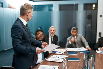 Confident mature businessman in formal wear holding some documet and discussing something with multicultural team while having a meeting in the modern office