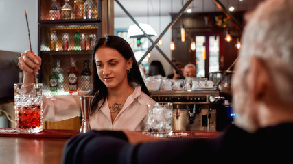 Tasty drink for every client. Young beautiful female bartender making a cocktail for man who visiting barbershop
