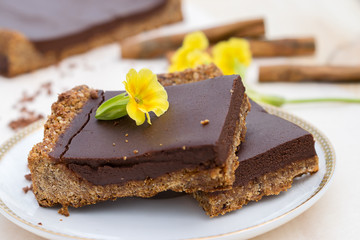 Сhocolate tart made from oatmeal, coconut milk and oil, dates and almonds on a white saucer.Cinnamon sticks and yellow flowers in the background.Сoncept of healthy food and healthy dessert