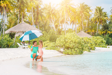 Young father and his little daughter walking under umbrella on white sand beach
