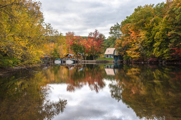 Fototapeta na wymiar Boathouses and moored boats along a river with wooded banks on a cloudy autumn day. Stunning autumn colours and reflection in water. Lakes region, NH, USA.