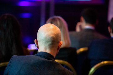 A man in a dark suit with an audio headset is sitting at a business conference.