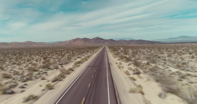 AERIAL - A drone flies straight over a long and empty road in the middle of the desert on a sunny day with tumbleweed next to the road