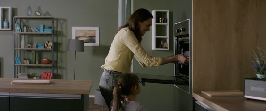 Mother and daughter cooking together at home, taking out bakery from the oven in the kitchen