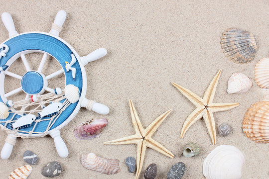Summertime vacation concept with rudder, different seashells, starfishes and stones on the sand. Travel and tourism. Top view. Copy space..