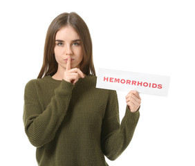Young woman holding paper with text HEMORRHOIDS and showing silence gesture on white background